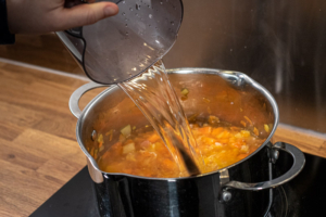 Water being poured into a saucepan containing bacon, carrot and onion